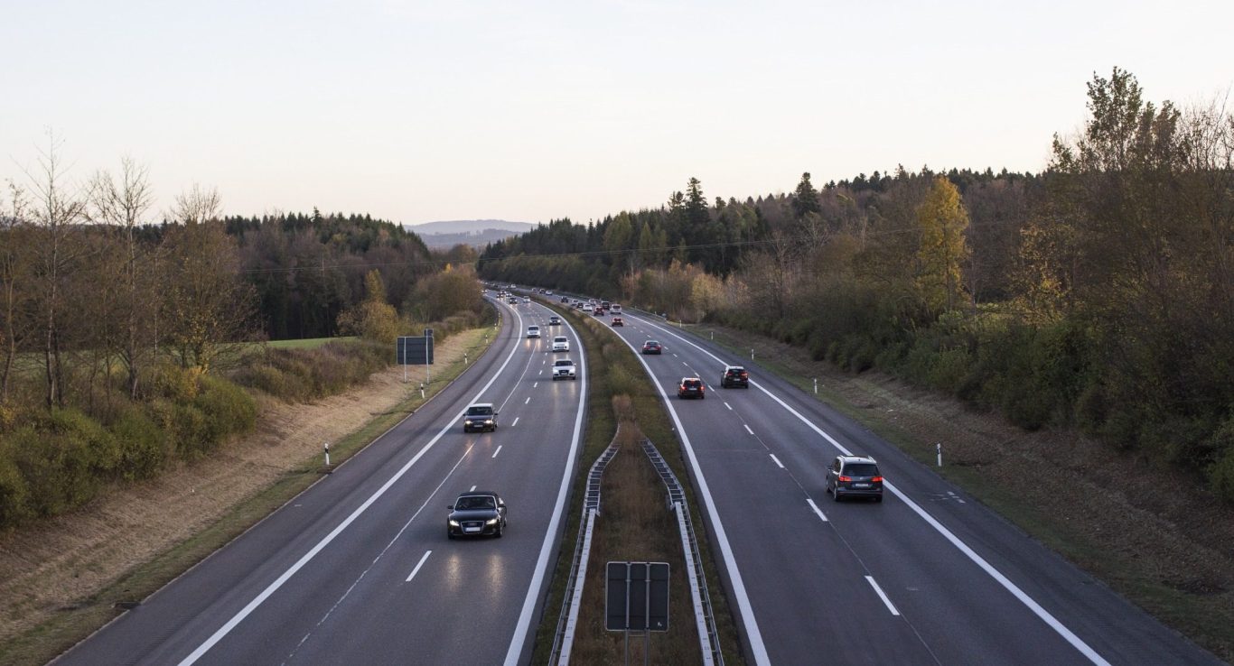 Blick auf eine zweispurige Autobahn in herbstlicher Landschaft. Mehrere Fahrzeuge fahren in beide Richtungen, umgeben von Wald und sanften H
