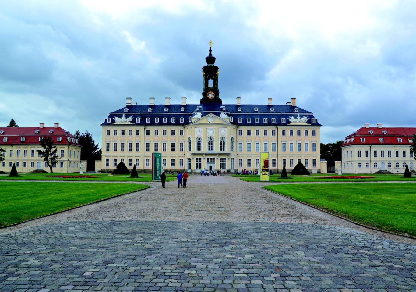 Schloss mit auffälligem Turm, umgeben von grünem Rasen und Wolken am Himmel.