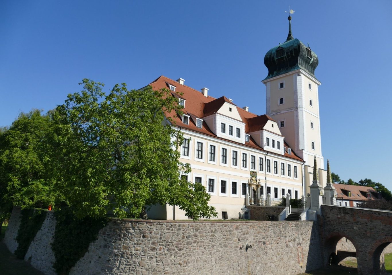 Historisches Schloss mit grünem Turm, umgeben von Bäumen und klarem blauem Himmel.