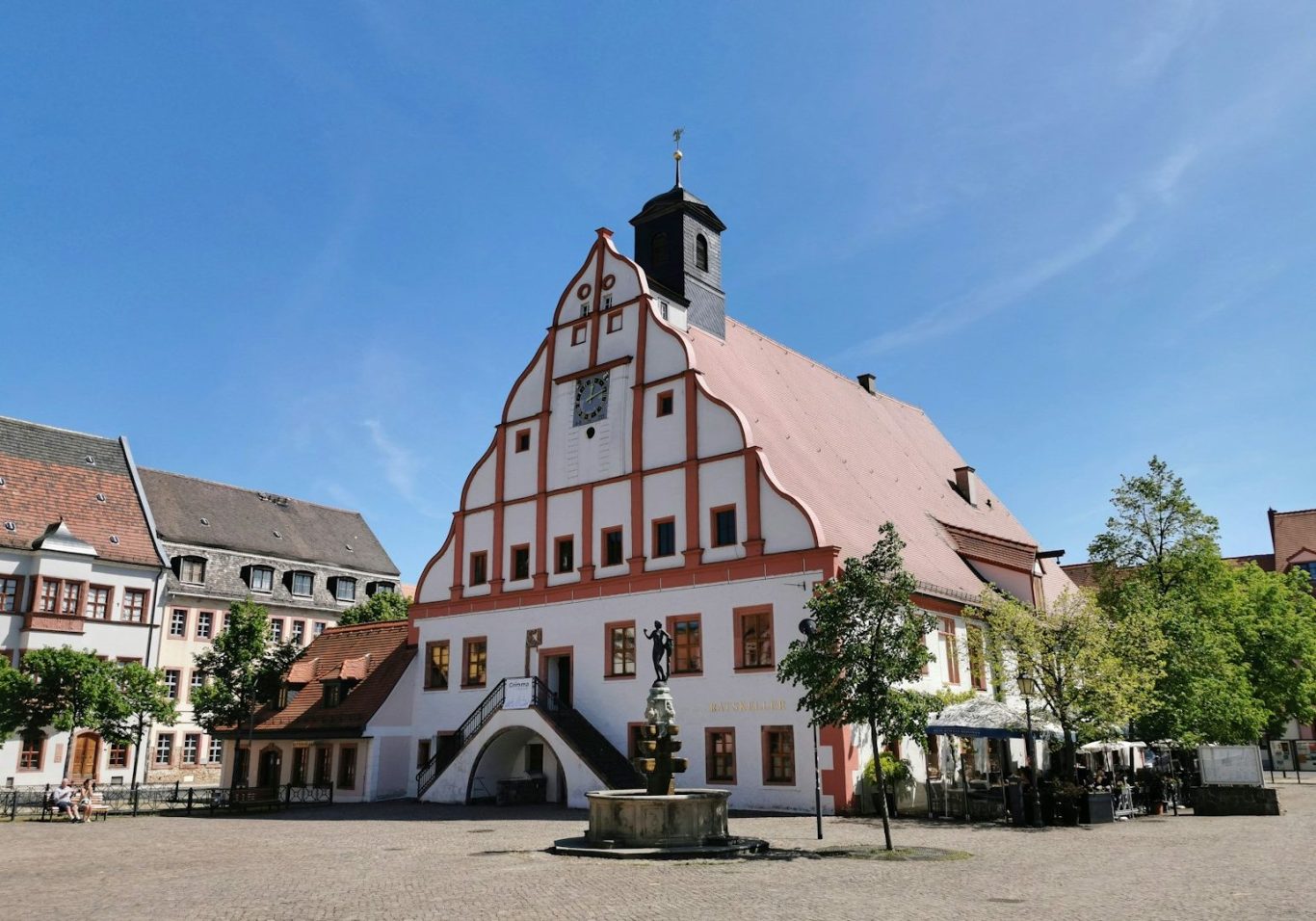 Fachwerkhaus mit rotem Dach und Turm, umgeben von Bäumen und einem Platz. Rathaus Grimma