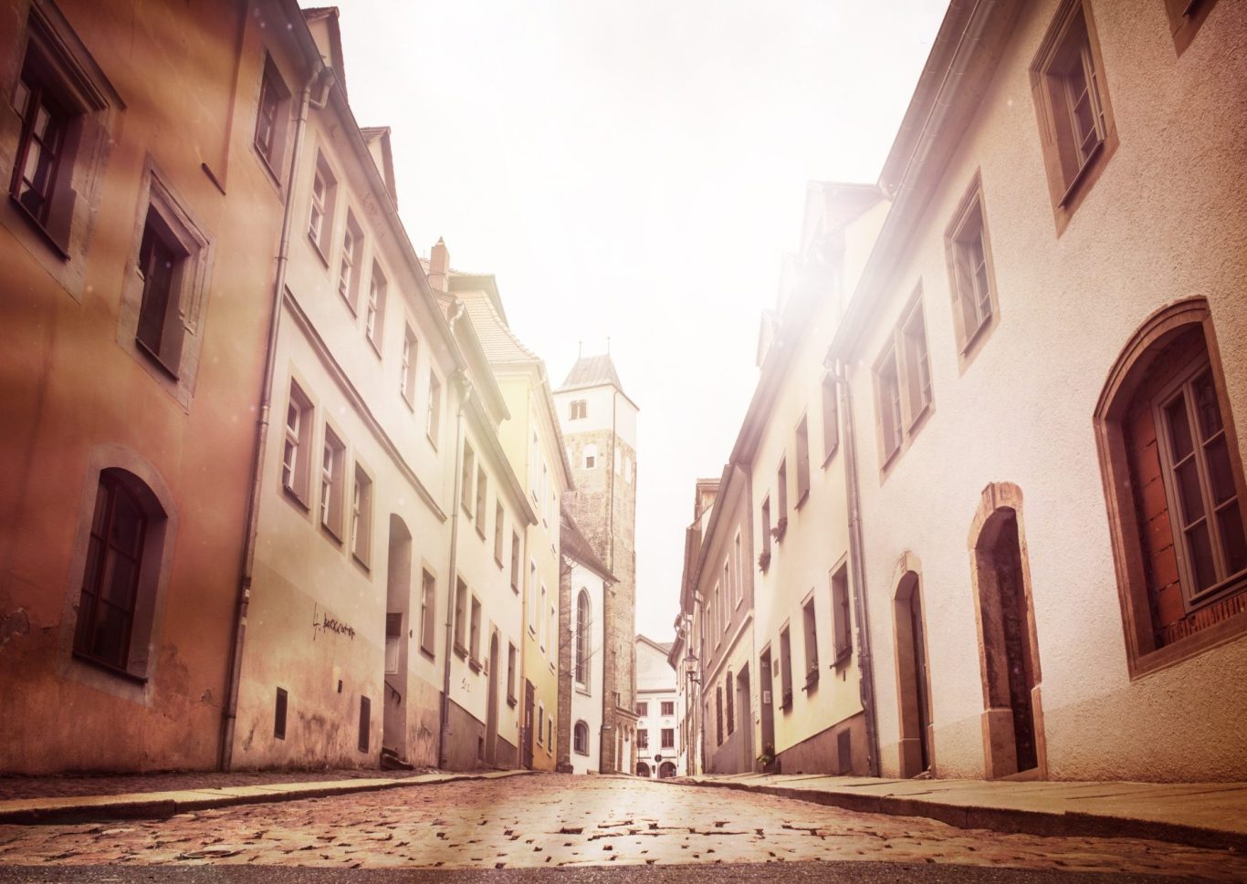 Enge, sonnige Straße mit historischen Gebäuden und einem Turm im Hintergrund.