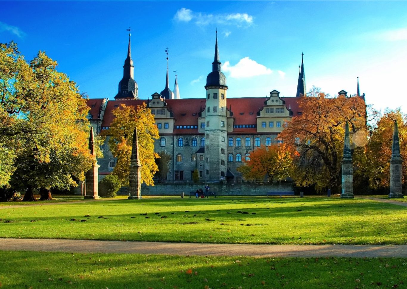 Schloss mit mehreren Türmen, umgeben von buntem Laub und grünem Rasen.