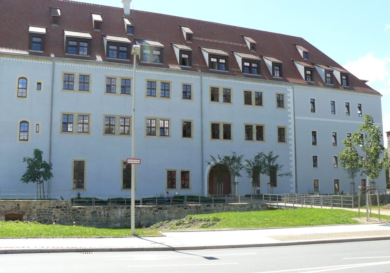 Helles Gebäude mit roten Dachziegeln und großen Fenstern, umgeben von Rasen und Bäumen.