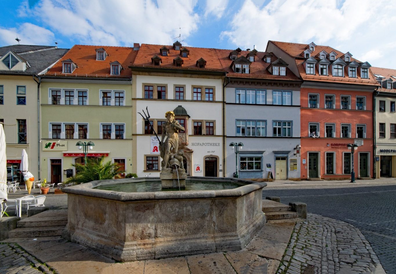 Brunnen in der Stadtmitte mit bunten Häusern im Hintergrund und blauen Himmel.