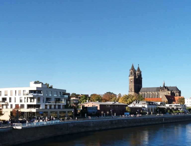 Sonnenbeschienenes Stadtbild von Magdeburg mit Kirche und modernen Gebäuden am Fluss.