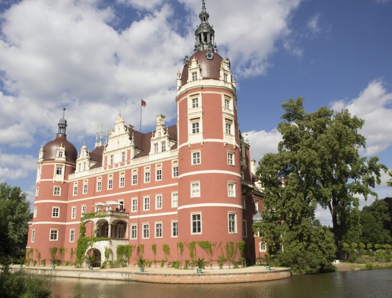 Schloss mit roter Fassade, Türmen und umgeben von Bäumen und Wasser.